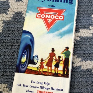 May include: Vintage travel brochure for Wyoming with Conoco. The brochure features a family standing near a classic car, with text that reads "For Long Trips Ask Your Conoco Mileage Merchant about TOURAIDE."