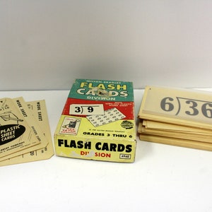 May include: A vintage Milton Bradley Flash Cards set for division, featuring a box with a green and red design and a stack of yellow cards with the equation "6)36" printed on them. The box includes a wipe-off plastic work sheet for test cards.