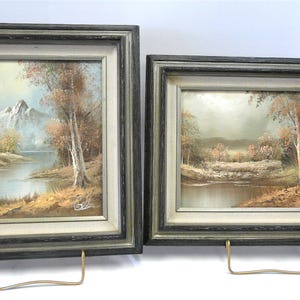 May include: Two framed landscape paintings. The paintings depict serene scenes with water, trees, and mountains. The frames are dark gray with a lighter inner border. The paintings are displayed on gold-colored stands.
