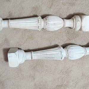 May include: Two antique, white-painted wooden balusters with ornate, classical designs. Each baluster features a square base, a fluted column, and a bulbous center section. The pieces are likely for architectural restoration.