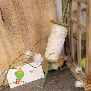 May include: A green wooden spool with white string wrapped around it. The spool is leaning against a wooden lattice. A white card with the word "kite" and a drawing of a kite is on the ground.
