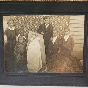 May include: A vintage black and white photograph of a family portrait. The photo features six people, including a baby, in a formal setting. The photograph is in a black frame.