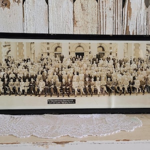 May include: A framed black and white photograph of a large group of people, likely a historical gathering. The photograph is in a black frame and features a building in the background. The text at the bottom of the image is partially visible.
