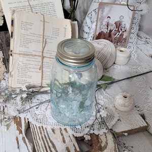 May include: A vintage blue glass Atlas mason jar with a silver lid. The jar is surrounded by antique books, twine, and a sepia-toned photograph, creating a rustic, nostalgic aesthetic. The jar is empty, and the word "Atlas" is embossed on the front.