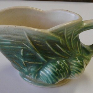 May include: A green and white ceramic creamer with a pine cone design. The creamer has a handle and a rounded base.