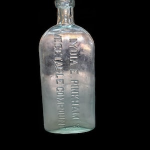 May include: A clear, antique glass bottle with the embossed text "LYDIA E. PINKHAM'S VEGETABLE COMPOUND" in a vertical arrangement. The bottle has a rounded body and a narrow neck, with a slightly bluish tint.