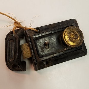May include: A dark brown, rectangular metal rim lock featuring a brass-colored circular knob with a small embossed floral emblem. The lock mechanism includes a protruding brass bolt and a separate matching strike plate, with a piece of twine attached.
