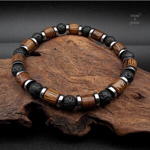 Könnte beinhalten: Ein Armband mit abwechselnd dunklen Lavasteinperlen und holzfarbenen, fassförmigen Perlen mit silberfarbenen Metallakzenten. Die Holzperlen haben ein gestreiftes Muster. Das Armband wird auf einem Stück dunklem Holz präsentiert.