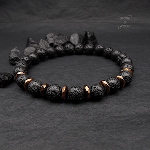 Op de afbeelding: Een zwarte lavasteen armband met goudkleurige accenten. De armband heeft ronde, getextureerde zwarte kralen afgewisseld met facetgeslepen goudkleurige kralen. De armband wordt gepresenteerd op een donkere ondergrond, met extra zwarte stenen op de achtergrond. Het logo "STONE & WOOD" is zichtbaar.