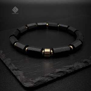 Könnte beinhalten: Ein Armband bestehend aus zylindrischen schwarzen Perlen und kleinen goldfarbenen Metallzwischenstücken in abwechselnder Anordnung. Die Perlen haben eine matte, texturierte Oberfläche. Ein Logo mit dem Text STONE & WOOD ist oben links sichtbar.
