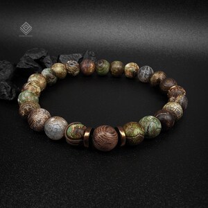 Könnte beinhalten: Ein elastisches Armband aus runden Perlen in Grün-, Braun- und Grautönen mit rissiger Textur. Das Design zeichnet sich durch eine zentrale Holzperle aus, die von zwei kupferfarbenen Metallabstandshaltern flankiert wird und einen Blickfang bildet.