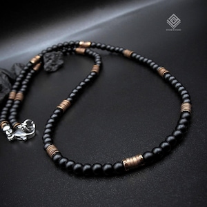 Könnte beinhalten: Eine Perlenkette mit mattschwarzen, kugelförmigen Perlen, die mit zylindrischen, kupferfarbenen Metallzwischenstücken durchsetzt sind. Die Kette hat einen silberfarbenen Karabinerverschluss und ein kleines Logo in der Ecke mit der Aufschrift STONE IS WOOD.