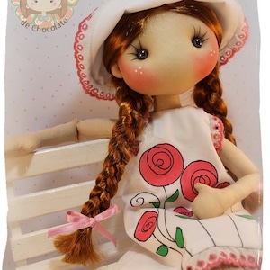 Puede incluir: Una muñeca hecha a mano con cabello rojo en trenzas, que lleva un vestido blanco con rosas rosas y un sombrero blanco con encaje rosa y flores rosas. La muñeca está sentada en un banco de madera blanco.