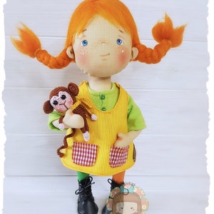 Puede incluir: Una muñeca hecha a mano con el pelo rojo en trenzas, que lleva un vestido amarillo con bolsillos a cuadros rojos y blancos, sosteniendo un mono de crochet marrón.