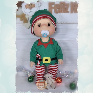 Puede incluir: Una muñeca de elfo hecha a mano que lleva un atuendo verde y rojo con un bastón de caramelo y un chupete. La muñeca está de pie sobre una superficie blanca con una guirlande blanca y plateada y adornos rojos y verdes.