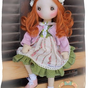 May include: A handmade doll with red hair, wearing a pink cardigan, a green and white floral dress, and white socks with pink shoes. The doll is sitting on a wooden surface and has a flower crown on her head. The doll is labelled "Pepitas de Chocolate".