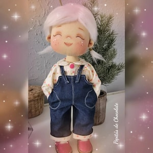 Puede incluir: Una muñeca hecha a mano con cabello blanco, que lleva una camisa blanca con estampado floral y un mono vaquero azul. La muñeca tiene mejillas rosadas y los ojos cerrados, y lleva zapatos rosas.