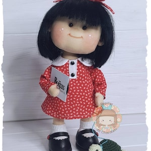 Puede incluir: Una muñeca hecha a mano con cabello negro, que lleva un vestido rojo con lunares blancos, zapatos negros y un lazo rojo. La muñeca sostiene un álbum de discos con el logotipo de los Beatles. La muñeca está de pie junto a una tortuga de crochet verde.