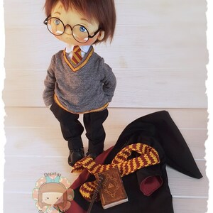May include: A handmade doll dressed as a wizard, wearing a grey sweater with a red and gold tie, black trousers, and round glasses. The doll is standing next to a black robe with a red lining, a gold and brown scarf, a book, and a wand.