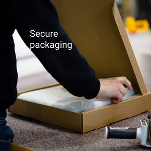 A person is opening a cardboard box with a hand, and there is a roll of tape and a box cutter on the table.