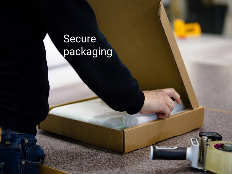 A person is opening a cardboard box with a hand, and there is a roll of tape and a box cutter on the table.