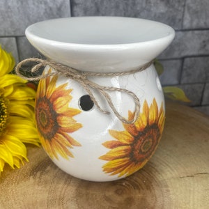 May include: White ceramic wax melt burner featuring a sunflower design. The burner has a wide, shallow top for holding wax melts and a smaller, round bottom. The sunflower design is in shades of yellow and brown.