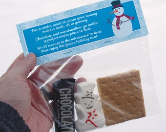 Op de afbeelding: Een doorzichtige plastic zak gevuld met een chocolade reep, een marshmallow sneeuwpop en een graham cracker. De zak heeft een blauw label met een sneeuwpop afbeelding en tekst die luidt: "Voor een wintersnack om je buik te warmen, maak een stapel, oh zo lekker. Chocolade en marshmallow gaan erin, een perfecte winterplek om te verstoppen. 10-15 seconden in de magnetron om te verwarmen, geniet dan van deze feestelijke traktatie."