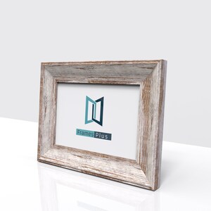 May include: A rustic wood picture frame with a whitewashed finish. The frame has a simple, rectangular design with a slightly raised edge. The frame is empty and ready to display a photo or artwork. The frame is made by Frames Plus.