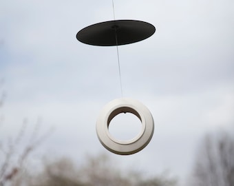May include: A white bird feeder with a black metal top hanging from a tree branch. The feeder is shaped like a ring with a hole in the center.