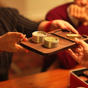 May include: A wooden tray with two silver metal grinders, one filled with green herb, and a small gold rectangle. The tray is being passed from one person to another.