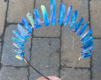 May include: A handcrafted crystal crown featuring iridescent blue crystals. The crystals are arranged in an arc and attached to a thin, gold-colored metal band with copper wire. The crown is designed for special occasions.