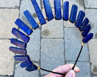May include: A blue lapis lazuli stone headband with gold wire accents. The headband is made of a black elastic band.