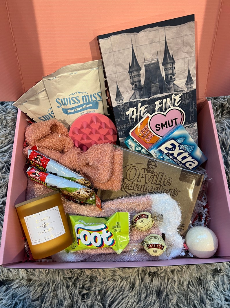 Op de afbeelding: Een roze cadeaudoos gevuld met verschillende items, waaronder een notitieboekje met de tekst "The Fine Smut", een roze badbom, een kaars, een pak Swiss Miss warme chocolademelk, een pak Orville Redenbacher's popcorn, een pak Fruit by the Foot snacks en een pak Bailey's Irish Cream chocolaatjes.