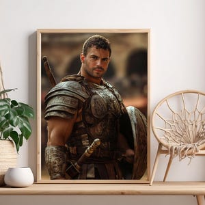 May include: A framed print of a man in ancient Roman armor, holding a shield and sword. The artwork is displayed on a wooden table, alongside a potted plant in a woven basket and a small white ceramic pot. A macrame plant hanger and a woven chair are also visible.