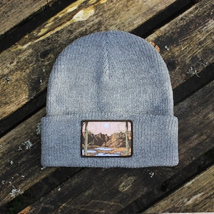 May include: Gray knitted beanie with a brown and green embroidered patch depicting a landscape with a lake and mountains.