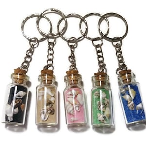 May include: Five glass bottle keychains filled with colorful sand and seashells. Each bottle has a cork stopper and is attached to a silver chain and keyring. The sand colors include black, tan, pink, green, and blue.