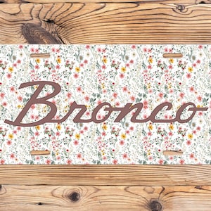 May include: A license plate with a floral pattern of pink, yellow, and green flowers on a white background. The word "Bronco" is in a brown script font across the center of the plate. The plate has four mounting holes.