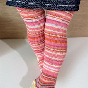 May include: Close-up of doll legs wearing pink and orange striped socks under a denim skirt. The socks have horizontal stripes in shades of pink, orange, yellow, and purple.