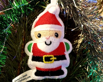 Puede incluir: Adorno bordado de Papá Noel para el árbol de Navidad. El adorno presenta un gorro y traje rojos con ribetes blancos, un cinturón negro con hebilla dorada y detalles verdes. Cuelga de un árbol de Navidad.