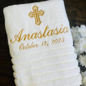 May include: White embroidered towel with the name "Anastasia", a gold cross, and the date "October 12, 2025". The towel has a ribbed texture and is likely a baptism or christening gift.