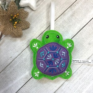 May include: Green felt turtle ornament with a purple shell decorated with a blue and white geometric pattern. The ornament has a white hanging loop and a tag that reads "Handmade in Canada by Gift of Embroidery.com".