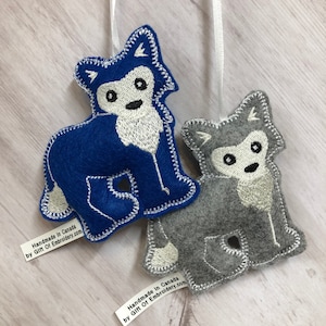 May include: Two felt wolf ornaments. One is blue, the other is gray. Both have white embroidered details, including the face, chest, and paws. Each has a white ribbon for hanging. The ornaments are handmade.