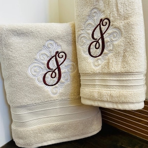 May include: Two cream-coloured towels with embroidered white and burgundy floral designs featuring the letter "J" in the centre.