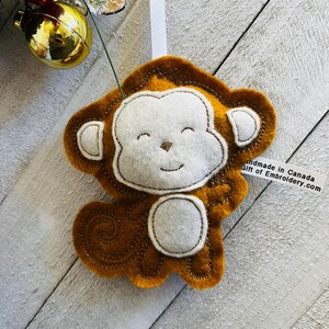 May include: A felt monkey ornament with a smiling face. The monkey is brown with a white face and a white tail. The ornament is handmade in Canada.