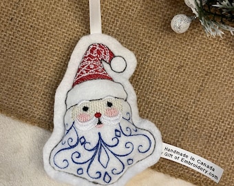 Puede incluir: Adorno navideño de fieltro blanco con la forma de Papá Noel, con un gorro rojo y blanco estampado y una barba blanca con detalles bordados en azul. Tiene una cinta blanca para colgar y una etiqueta que dice "Handmade in Canada / Gift of Embroidery.com".