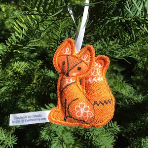 May include: Orange felt fox ornament with white embroidered details, hanging from a white ribbon. Handmade in Canada by Gift of Embroidery.com