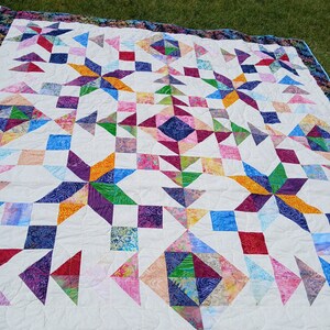May include: A colorful quilt with a geometric pattern of squares, triangles, and stars. The quilt is made with a variety of fabrics in shades of blue, green, purple, pink, orange, and yellow. The quilt is edged with a dark blue and purple fabric with a floral pattern.