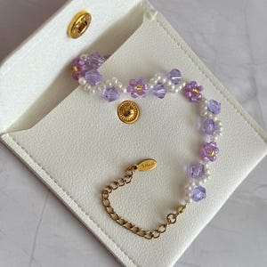 May include: A delicate bracelet featuring lavender and white beads, accented with gold-colored metal. The bracelet is displayed on a white, square, leather-like case with a gold snap closure. The clasp is marked with "S.steel".