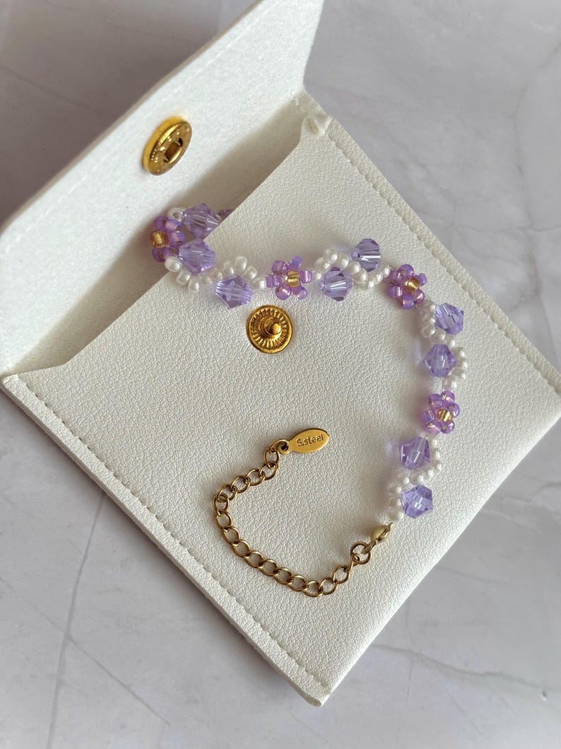May include: A delicate bracelet featuring lavender and white beads, accented with gold-colored metal. The bracelet is displayed on a white, square, leather-like case with a gold snap closure. The clasp is marked with "S.steel".