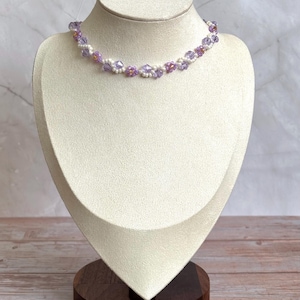 May include: A delicate necklace with a floral design, featuring lavender and white beads. The necklace is displayed on a cream-colored jewelry bust with a dark wooden base. The beads are arranged in a repeating pattern.